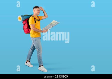 Happy millennial tourist with tourist gears and map looking ahead while walking on blue studio background, copy space Stock Photo