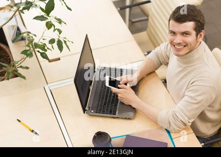 Top view of a handsome cheerful young man smiling to the camera using his smart phone working on the laptop at the loft office workspace copyspace stu Stock Photo