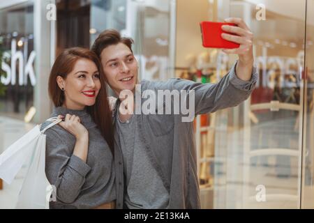 Adorable young loving couple using smart phone at the shopping mall, taking selfies together, copy space. Sharing, news, blogger concept Stock Photo