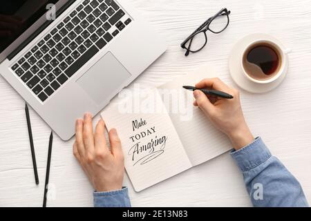 Man with cup of hot coffee writing text MAKE TODAY AMAZING in notebook  at table Stock Photo