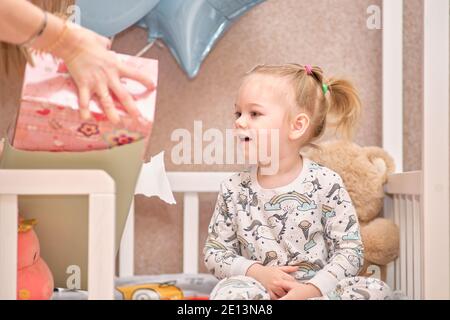 A little girl in pajamas receives birthday gifts while sitting in a white baby cot. Balloons in the background Stock Photo