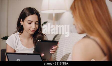 Charming caucasian friends with red and black hair sitting on the sofa and using tablet and laptop together Stock Photo