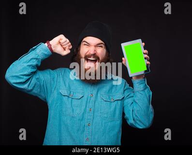 An exicted young bearded man is looking at the camera and is holding a tablet with green screen . Stock Photo