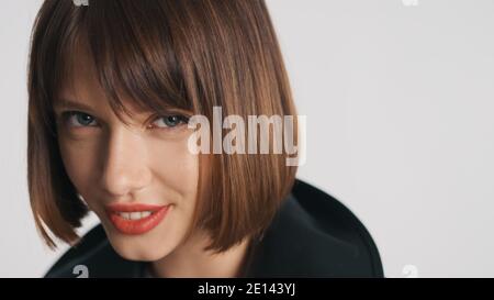 Close up beautiful blue eyed girl with bob hair and red lips slyly looking in camera and smiling over white background Stock Photo