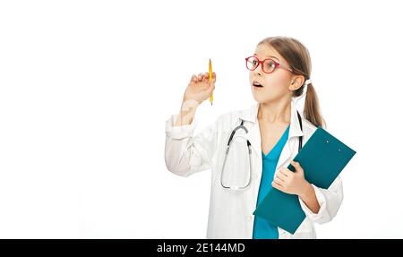 Cute girl wearing doctor's coat and stethoscope, with surprise emotions on her face, pointing pen into empty space on background. Stock Photo