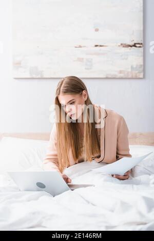 young freelancer in silk robe using laptop and holding papers in bedroom Stock Photo