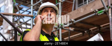 Panorama of male builder foreman, worker or architect on construction site holding building plans and talking on his cell phone panoramic web banner Stock Photo