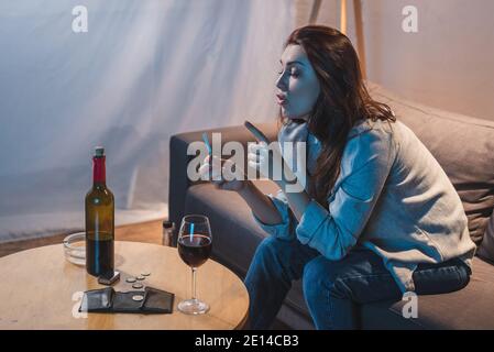 drunk woman holding cigarette while sitting near red wine, coins and empty wallet on table Stock Photo