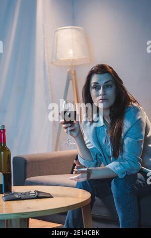 frustrated woman holding cigarette and glass of red wine near bottle and empty wallet on table Stock Photo