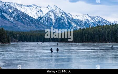 Tourists doing ice-skating in Johnson Lake frozen water surface in winter time. Snow-covered mountain in the background. Banff National Park Stock Photo