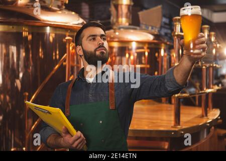 Bearded professional brewer holding clipboard, examining freshly made beer in a glass. Beer craftsman working at his microbrewery holding up beer mug. Stock Photo