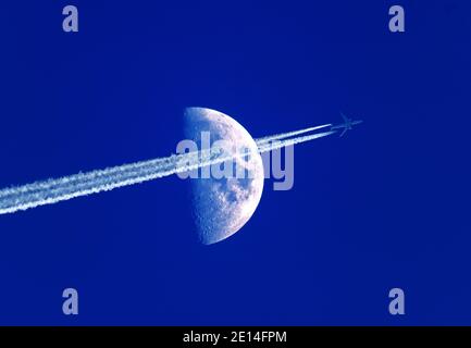 High flying jet aircraft with contrails passing in front of a half moon at dusk Stock Photo