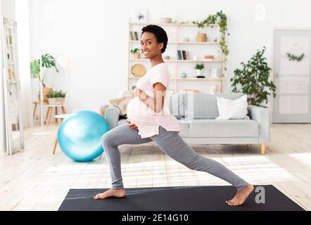 Active and sportive pregnancy. Cheerful pregnant black woman doing lunges, stretching her legs, exercising at home Stock Photo