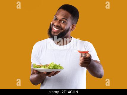 Cheerful Black Man Eating Veggie Salad Holding Plate, Yellow Background Stock Photo