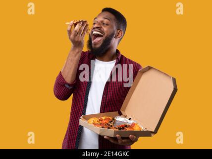 Black Man Eating Pizza Standing Holding Pizzeria Box, Yellow Background Stock Photo
