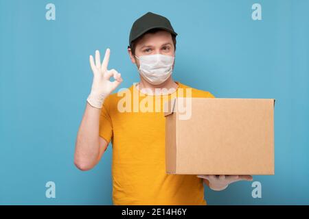 Delivery man holding cardboard boxes in medical rubber gloves and mask showing ok gesture. Studio shot on blue wall. Stock Photo