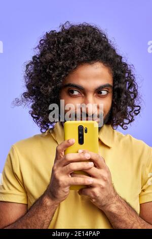 sly arabic male using smartphone, thinking what to write in message, looking at side, isolated over purple background Stock Photo