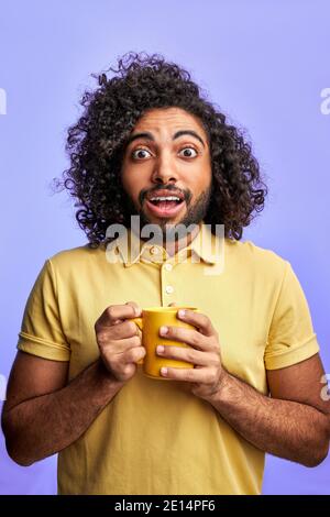 happy young arabic male with cup in hands, isolated over purple background. handsome guy with curly hair look at camera with opened eyes, surprised Stock Photo