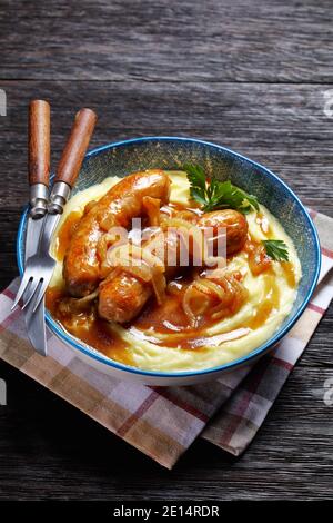 Bangers and mash traditional british dish of pork sausage with onion gravy and mashed potato on a blue plate with cutlery on a dark wooden background, Stock Photo