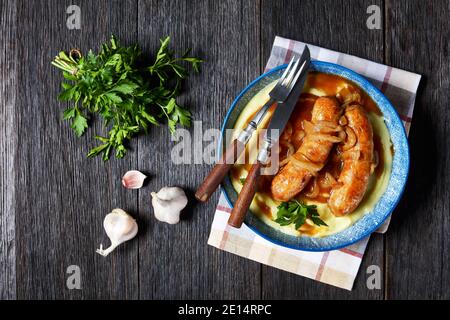 Bangers and mash traditional british dish of pork sausage with onion gravy and mashed potato on a blue plate with cutlery on a dark wooden background, Stock Photo