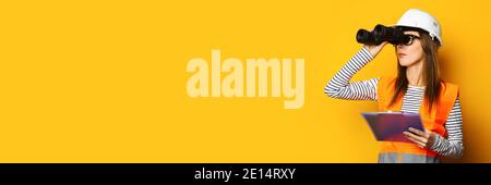 Young woman in signal vest and construction helmet looking through binoculars on yellow background. Stock Photo