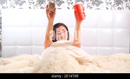 woman rises in the bed with an alarm clock and takeaway coffee in her hands, enjoys the morning and drinks coffee. The concept of starting a new day Stock Photo