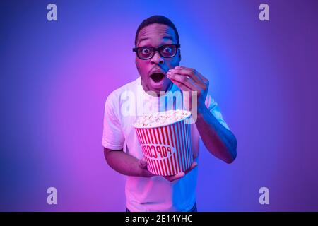 Shocked black guy in 3D glasses eating popcorn in cinema, neon light Stock Photo