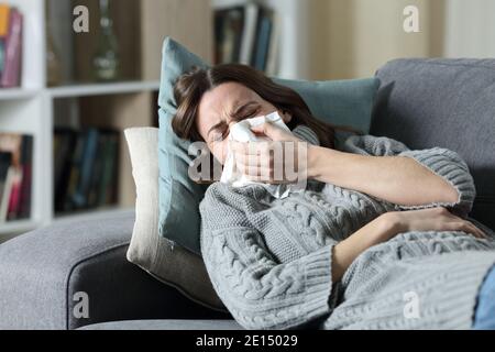 Ill woman suffering flu symptoms blowing on tissue lying on a couch at home