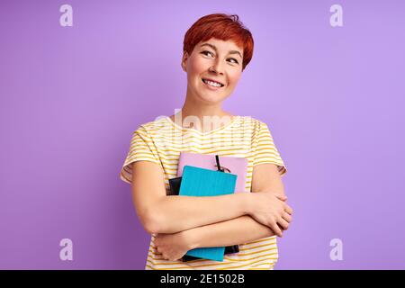 female holds books in hands andlooking at side, isolated over purple background. young redhead female enjoy studying, education Stock Photo