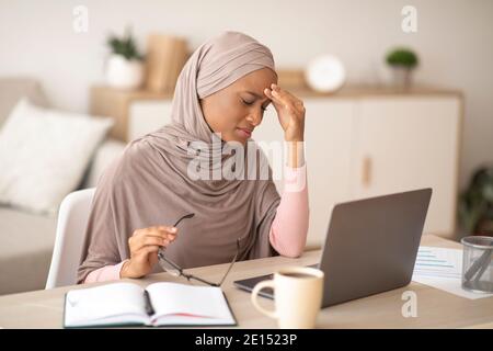 Deadline stress. Exhausted black woman in hijab tired after working on laptop for too long, having headache at home Stock Photo