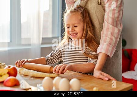 mother is teaching daughter to roll out dough for baking cookies, in the kitchen at home. on table Stock Photo