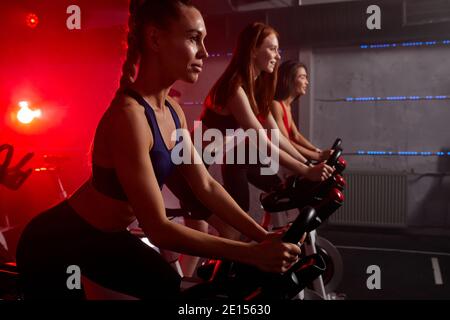 three women biking in gym, exercising legs doing cardio workout cycling bikes, spinning in health club, wearing tracksuit sportive outfit Stock Photo