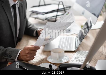 cropped view of trader holding papers with infographics near laptop and coffee cup Stock Photo