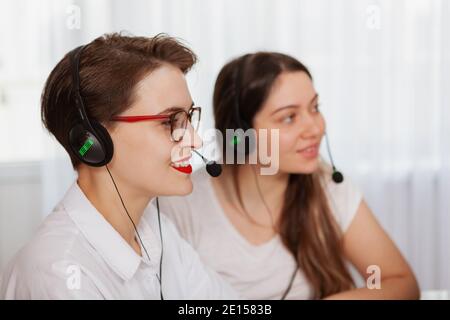 Two cheerful young women wearing headsets, working at call center. Customer support employees helping clients online, using headphones with microphone Stock Photo