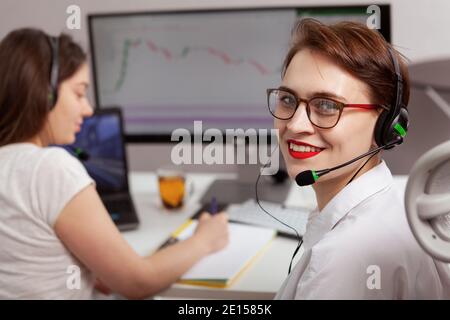 Cheerful young female customer support agent smiling to the camera, her colleague working on background Stock Photo