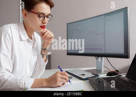Attractive young businesswoman working on two computers, writing notes on a paper Stock Photo