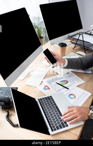 partial view of trader holding smartphone near laptop, computer monitors and infographics Stock Photo