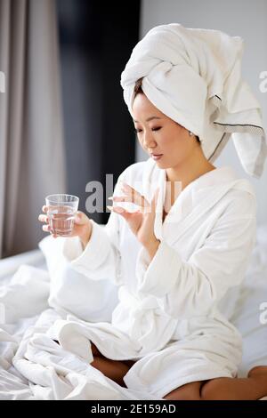 pregnant woman taking a medicine pill of vitamin while sitting on bed at home, wearing bathrobe and towel. early pregnancy, first month Stock Photo
