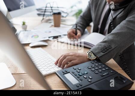 cropped view of trader talking on landline phone, writing in notebook and typing on laptop, blurred foreground Stock Photo