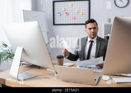 serious trader holding papers while sitting at workplace near laptop and monitors Stock Photo