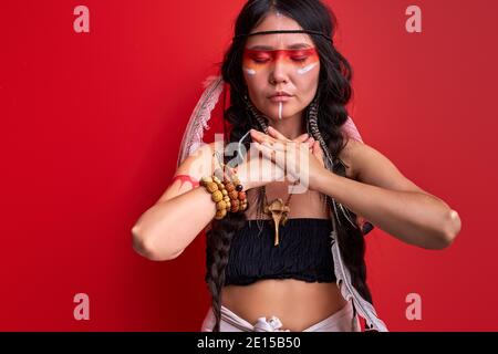tribal woman in shamanic clothes is shamaning with closed eyes, isolated over red background, portrait of ethnic woman with drawings on body Stock Photo