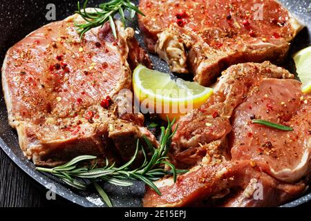 raw spiced pork chops with rosemary sprigs and lemon slices in a skillet on a wooden table, horizontal view from above, close-up Stock Photo