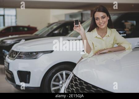 Young happy woman smiling joyfully, posing with a car key in her hand, leaning on her new automobile. Cheerful woman celebrating buying new car, copy Stock Photo