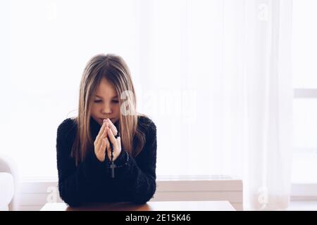 A cute girl prays in the living room with a rosary in hand. Stock Photo