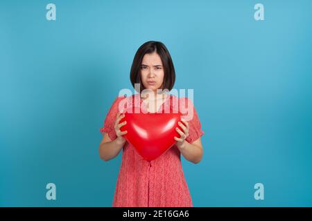 mock up offended by a bad gift, a young Asian woman holds a red heart-shaped balloon in her hands on Valentine's day, isolated on a blue background Stock Photo