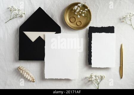 Elegant wedding stationery top view. Flat lay blank invitation cards mockup, golden decorations, gypsophila flowers on pastel white background. Stock Photo