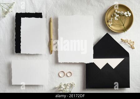 Wedding stationery still life scene top view. Flat lay invitations cards templates, black envelopes, golden pen, rings, flowers on textile background. Stock Photo