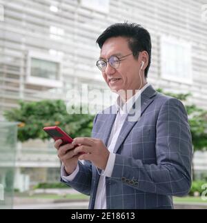 Asian businessman doing video call on smartphone using earphones in the street, smiling and looking at screen. Stock Photo