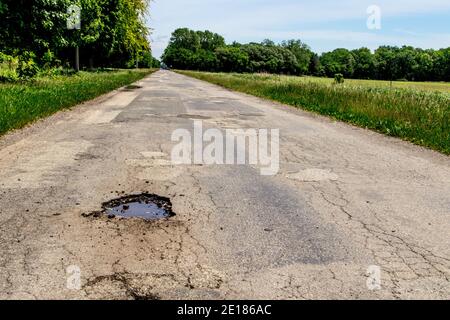 Terrible Michigan Roads. Rural two lane paved road with huge pothole, cracked aging asphalt and no lines painted on the pavement. Stock Photo