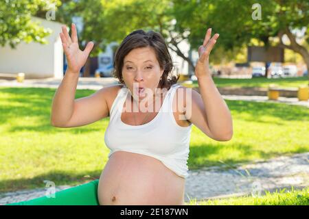 Frustrating pregnant lady. Photo of emotional angry screaming pregnant woman hands in air in frustration sitting outside, outdoors in a park on a gree Stock Photo